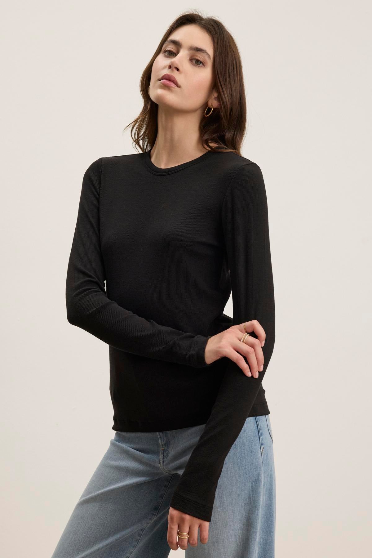   A person wearing the CAMINO TEE by Velvet by Jenny Graham, a black long-sleeve shirt crafted from soft modal rib fabric, pairs it with blue jeans against a plain background. This outfit highlights the top's slimmer fit, making it perfect for versatile layering. 