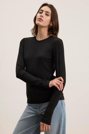 A person wearing the CAMINO TEE by Velvet by Jenny Graham, a black long-sleeve shirt crafted from soft modal rib fabric, pairs it with blue jeans against a plain background. This outfit highlights the top's slimmer fit, making it perfect for versatile layering.