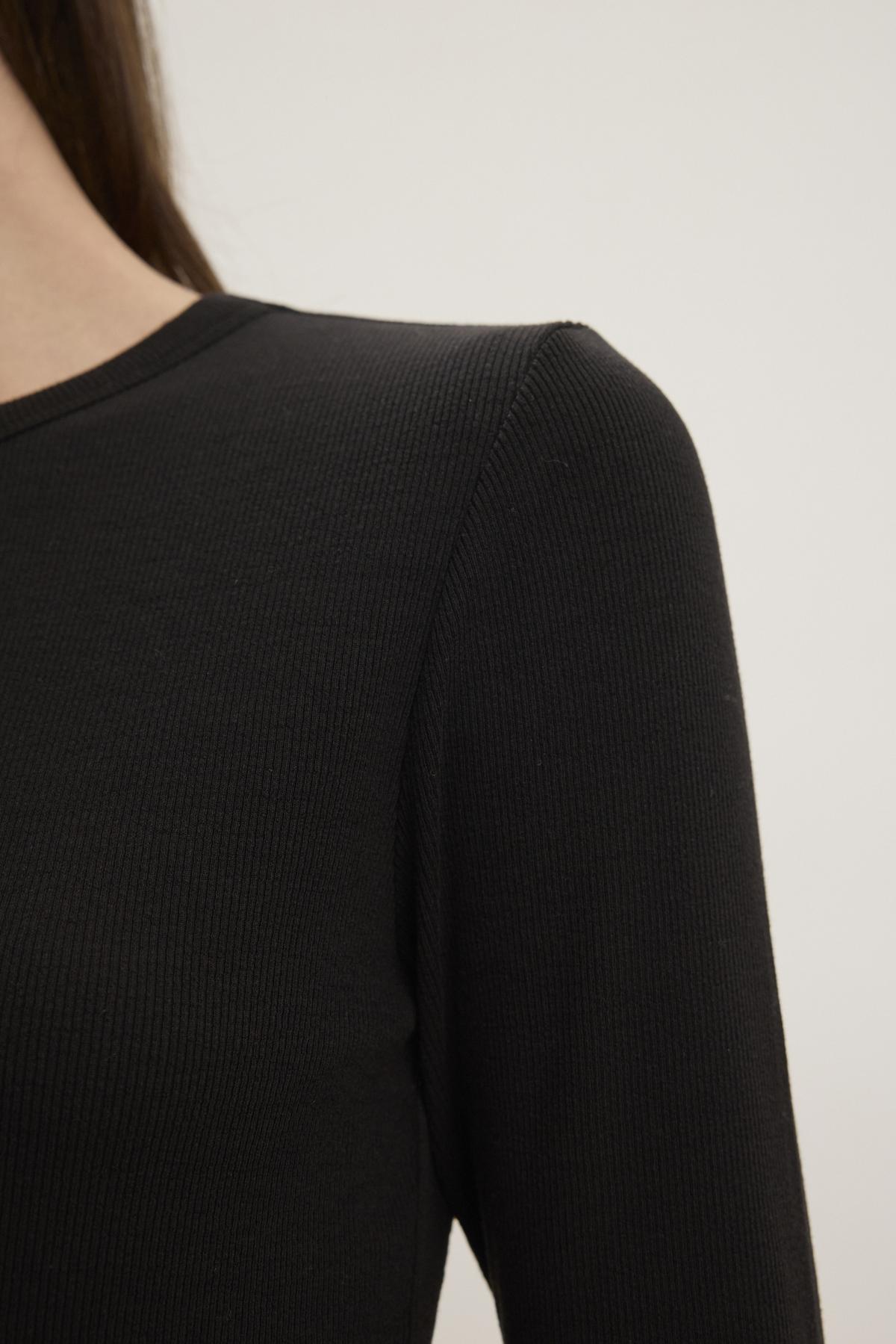   A close-up of a person wearing the Velvet by Jenny Graham CAMINO TEE, showcasing its sleek black modal rib material on the shoulder and part of the arm against a plain background. The slimmer fit enhances its timeless appeal. 