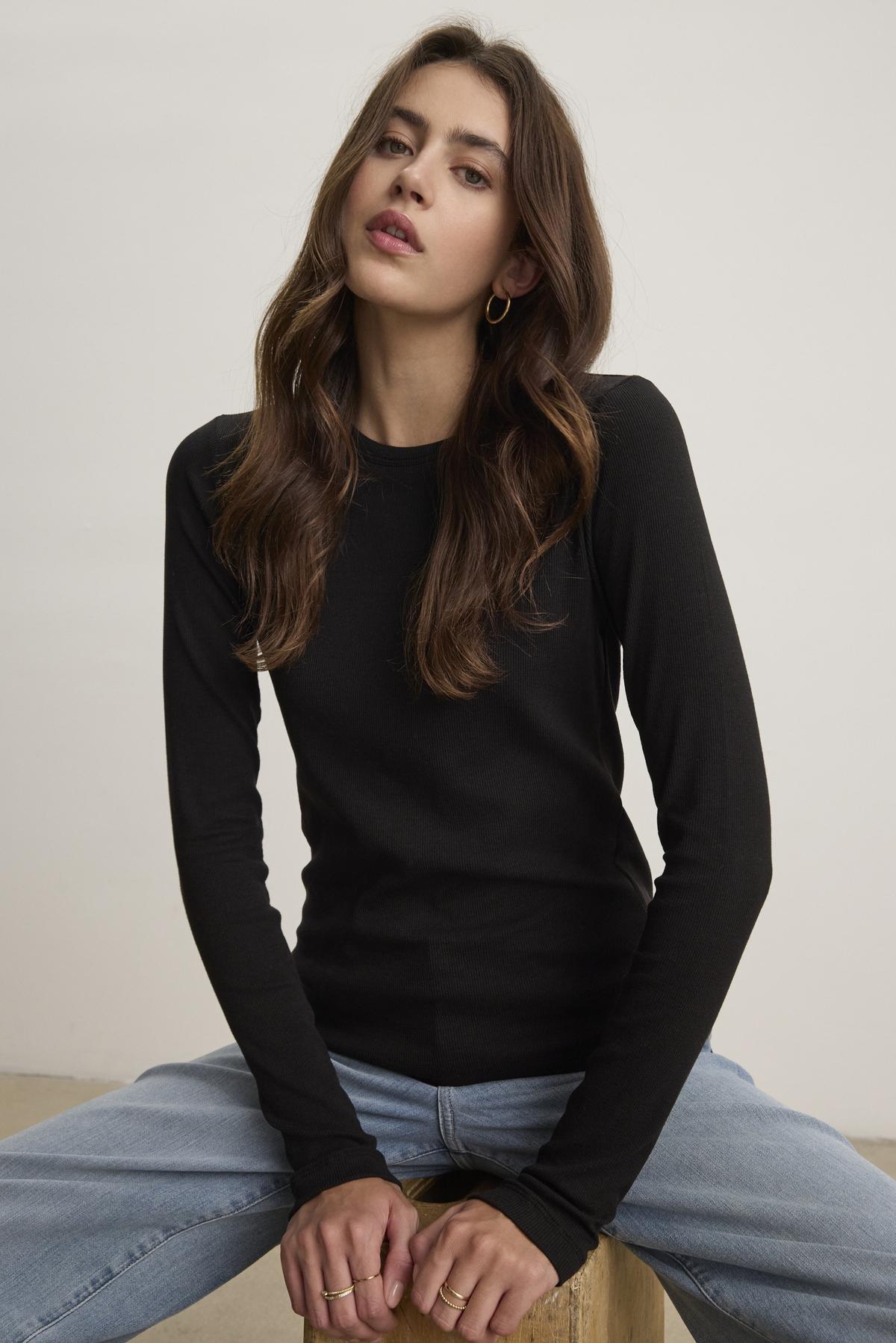   A person with long hair is wearing the black CAMINO TEE by Velvet by Jenny Graham, paired with blue jeans that have a slimmer fit, while sitting on a stool against a plain background. 