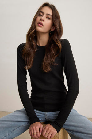 A person with long hair is wearing the black CAMINO TEE by Velvet by Jenny Graham, paired with blue jeans that have a slimmer fit, while sitting on a stool against a plain background.