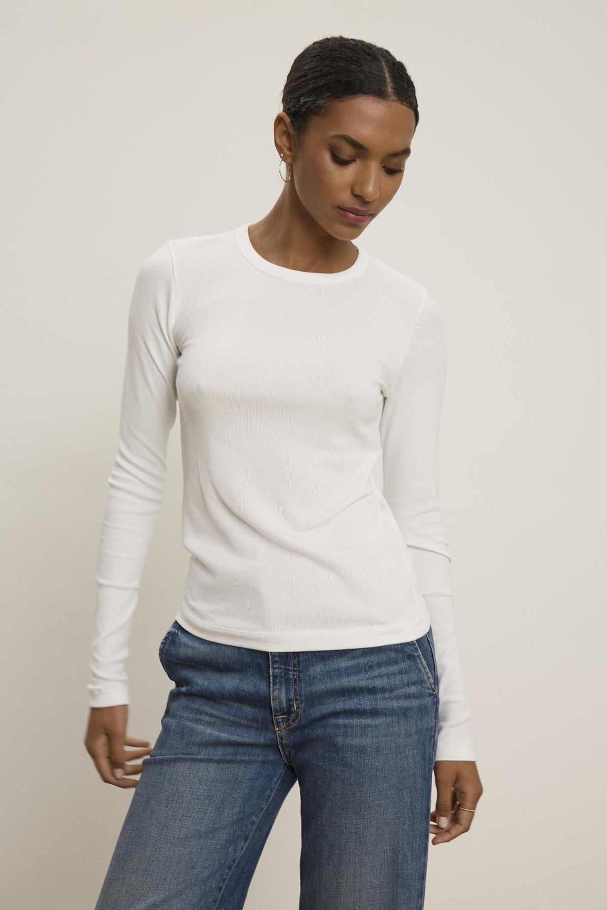   Clad in the elegant CAMINO TEE by Velvet by Jenny Graham paired with slimmer-fit blue jeans, a person stands against a plain background, showcasing a versatile style. 