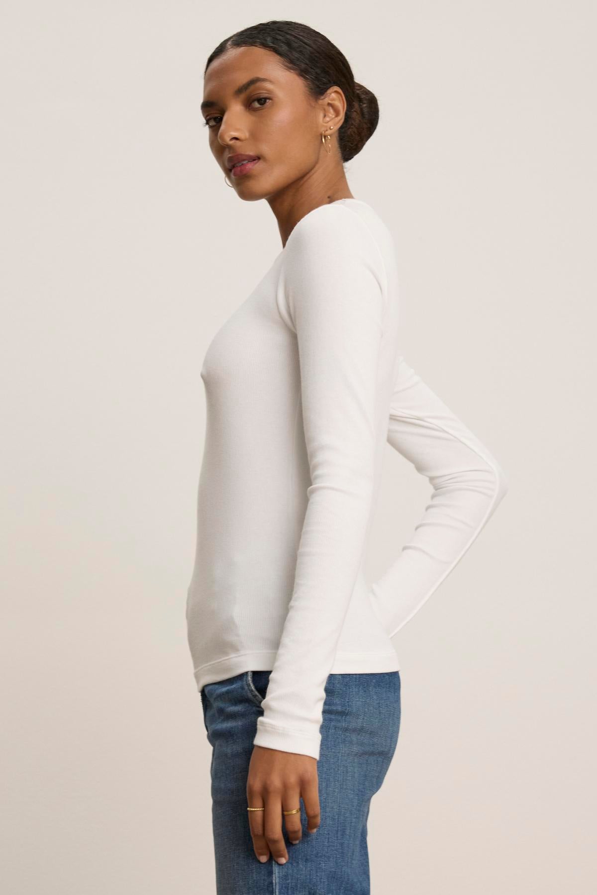   A person in the CAMINO TEE by Velvet by Jenny Graham and blue jeans stands sideways against a plain background, highlighting the timeless appeal and slimmer fit of this versatile outfit. 