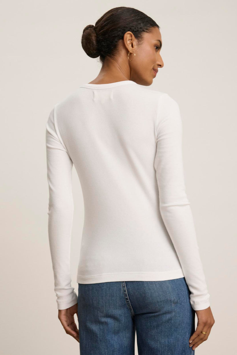 A person with tied-back hair is seen wearing the classic long-sleeved CAMINO TEE in white by Velvet by Jenny Graham, paired with a sleek pair of blue jeans, facing away from the camera.