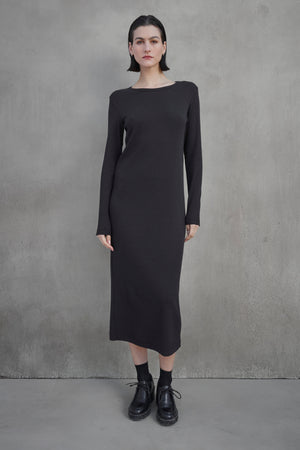 A person stands against a plain concrete wall, wearing the CORONADO DRESS by Velvet by Jenny Graham, which is a long-sleeved, ankle-length black dress, paired with black shoes.