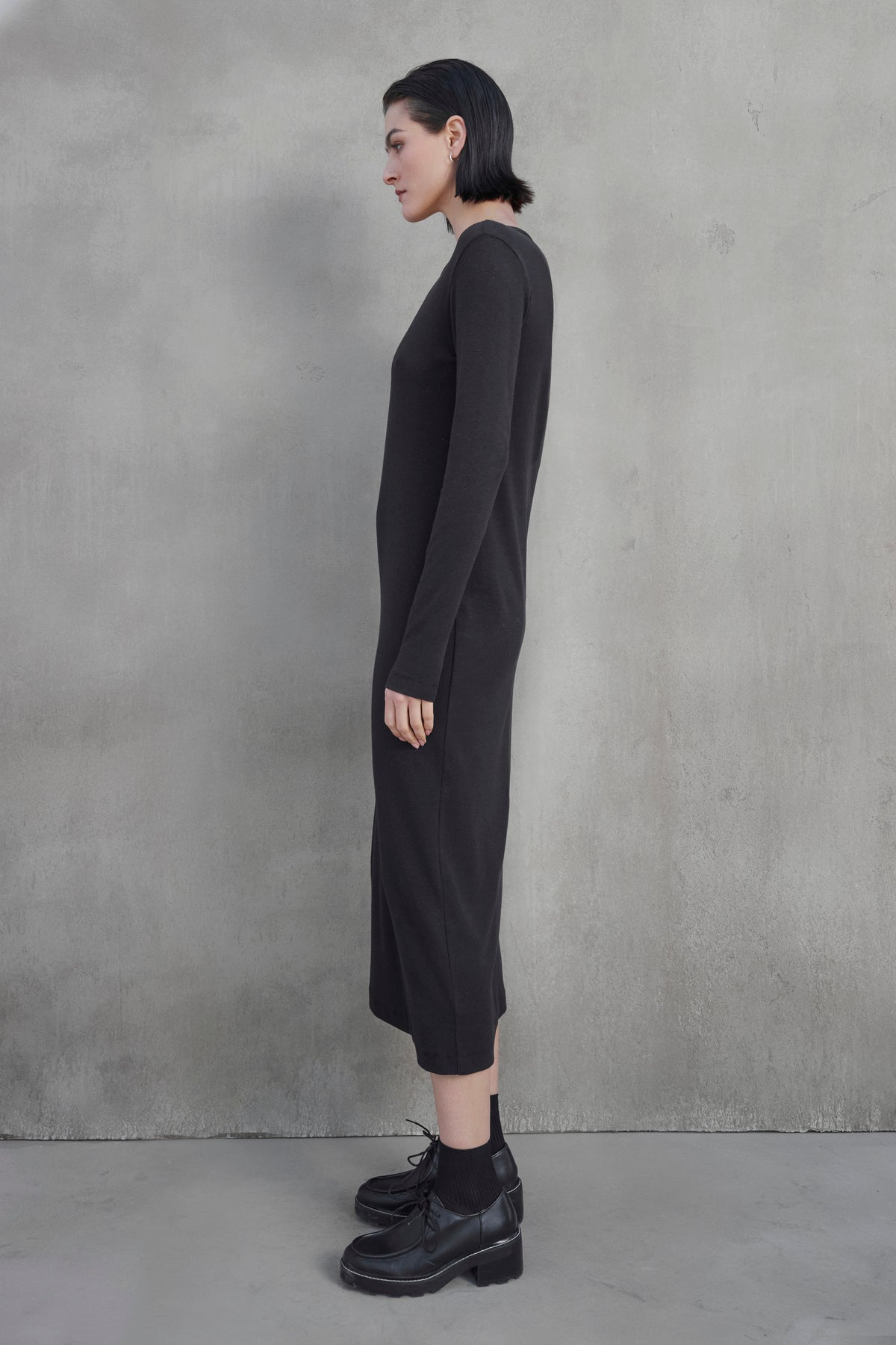   A person with short dark hair stands sideways against a gray textured wall, wearing a long black CORONADO DRESS by Velvet by Jenny Graham and black platform shoes. 