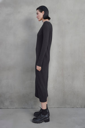 A person with short dark hair stands sideways against a gray textured wall, wearing a long black CORONADO DRESS by Velvet by Jenny Graham and black platform shoes.