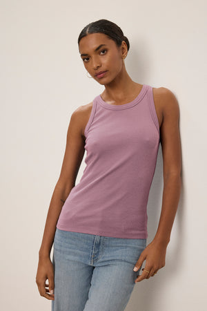 A person in the Velvet by Jenny Graham CRUZ TANK TOP, a sleeveless lavender piece made of ribbed cotton-modal for stretch, and blue jeans leans casually against the wall.