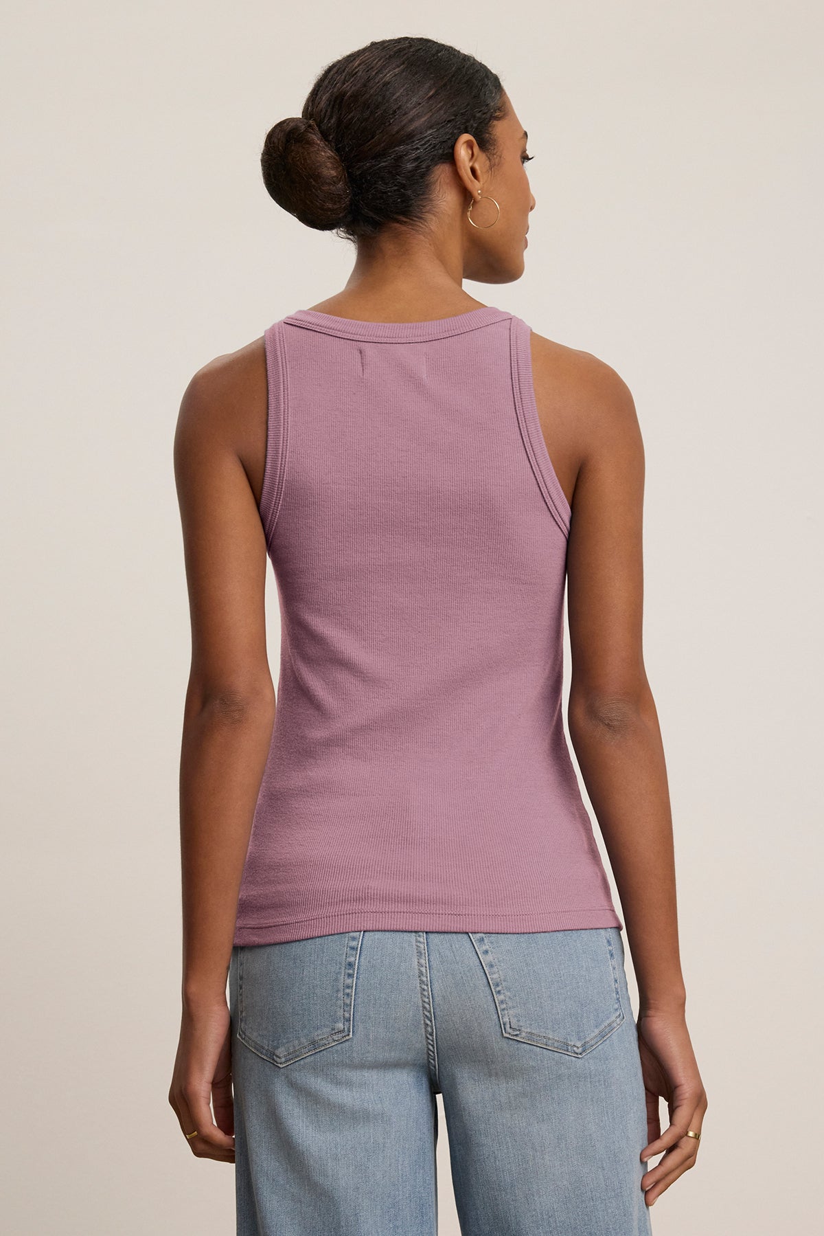   A person with their hair in a bun is wearing the Velvet by Jenny Graham CRUZ TANK TOP, a sleeveless purple ribbed cotton-modal top, paired with blue jeans, highlighting the versatility of a capsule wardrobe as they face away. 