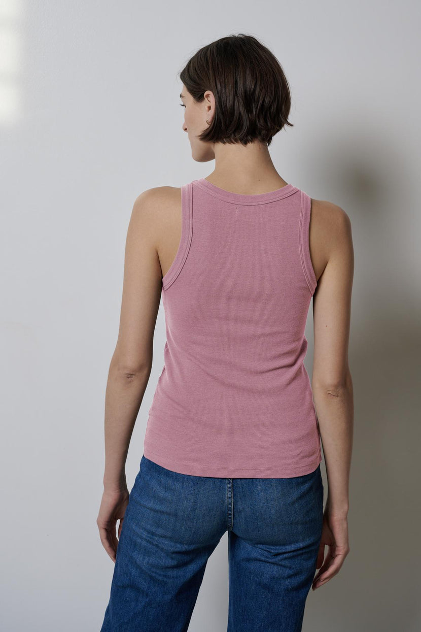 A person with short hair, standing against a plain wall in blue jeans, wears the Velvet by Jenny Graham CRUZ TANK TOP—a pink, versatile tank made from heavyweight ribbed cotton-modal blend—capturing the essence of a capsule wardrobe.