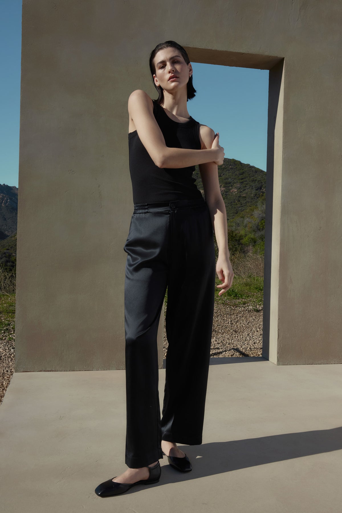   A person stands outdoors, leaning against a tall structure with an archway. They are wearing a black sleeveless top, high-waisted MODESTO SILK CHARMEUSE PANT by Velvet by Jenny Graham, and black shoes. Hills and blue sky are visible in the background. 