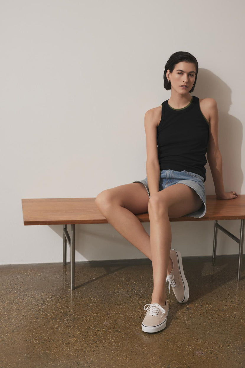 A person with short dark hair, dressed in a black CRUZ TANK TOP by Velvet by Jenny Graham, denim shorts, and white sneakers, sits on a wooden bench against a plain wall.