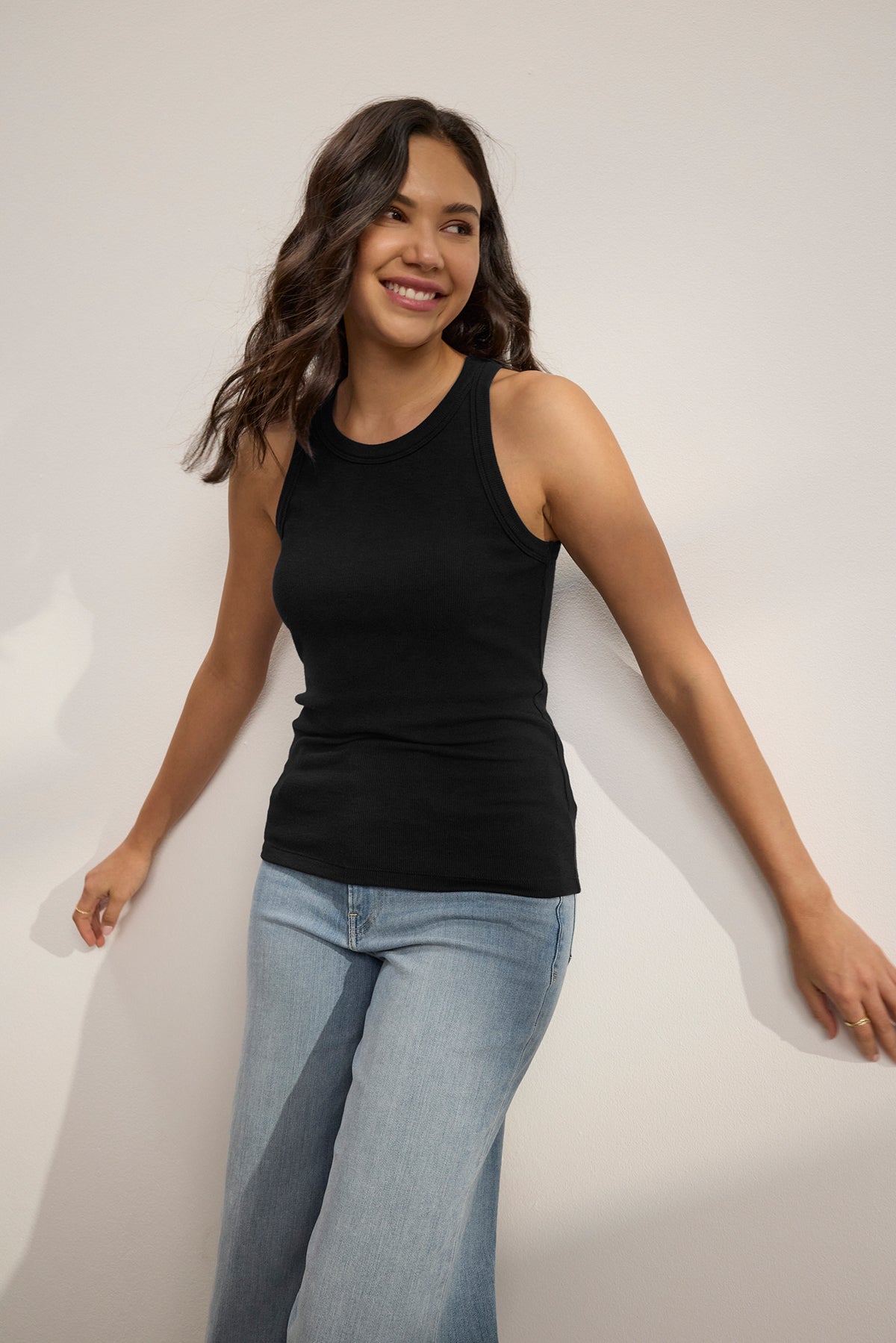   Wearing Velvet by Jenny Graham's CRUZ TANK TOP and blue jeans, a person stands against a white wall, smiling and looking to the side. 