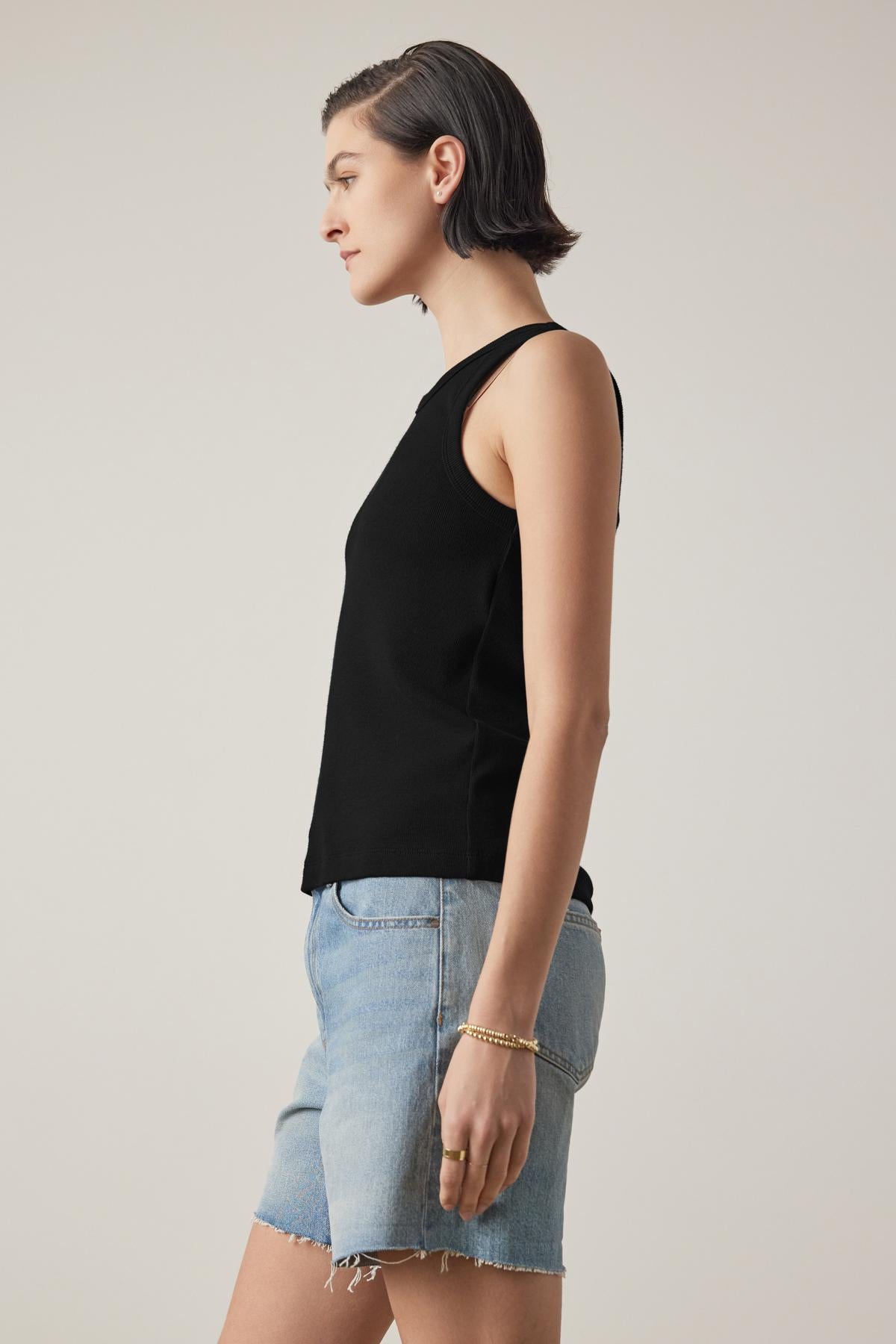   A woman with short dark hair is standing in a profile view, wearing the Velvet by Jenny Graham CRUZ TANK TOP, which is sleeveless and made from ribbed cotton-modal blend, paired with light denim shorts, against a plain background. 