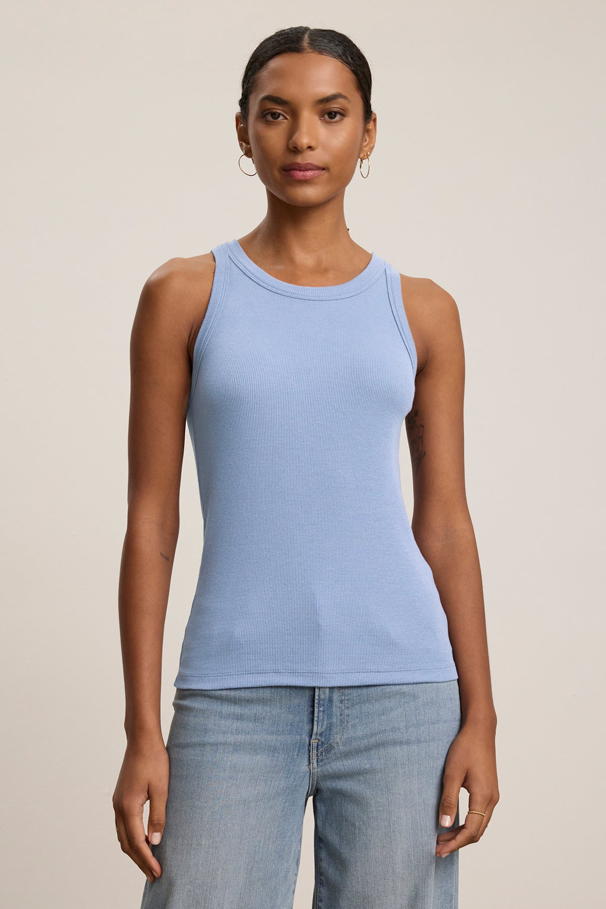   A person models the CRUZ TANK TOP in light blue by Velvet by Jenny Graham, designed from a ribbed cotton-modal blend. Paired with blue jeans against a neutral background, it highlights its essential role in any capsule wardrobe. 