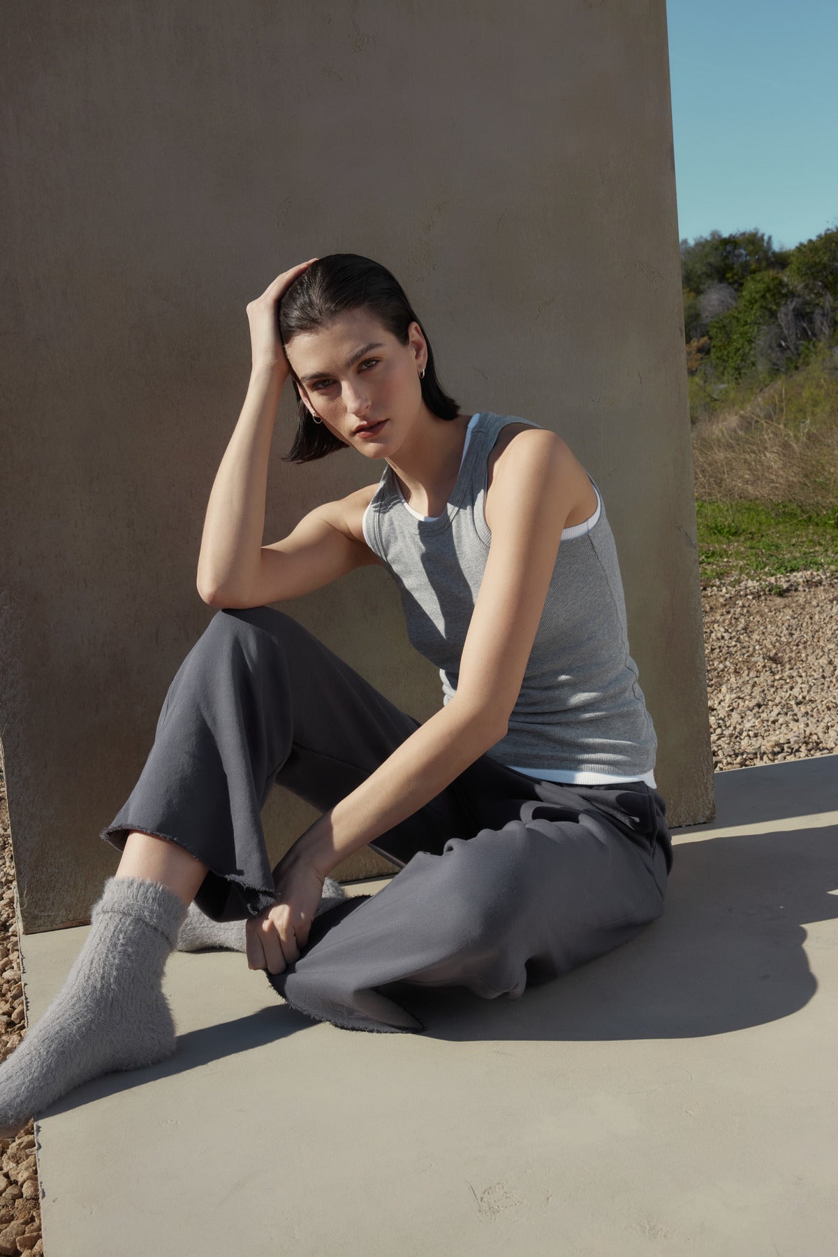   A person with short dark hair in a versatile tank and sweatpants sits on a concrete surface, resting their head on one hand, with grass and trees in the background. The ribbed cotton-modal blend of the Velvet by Jenny Graham CRUZ TANK TOP in gray adds a touch of effortless style, perfect for any capsule wardrobe. 