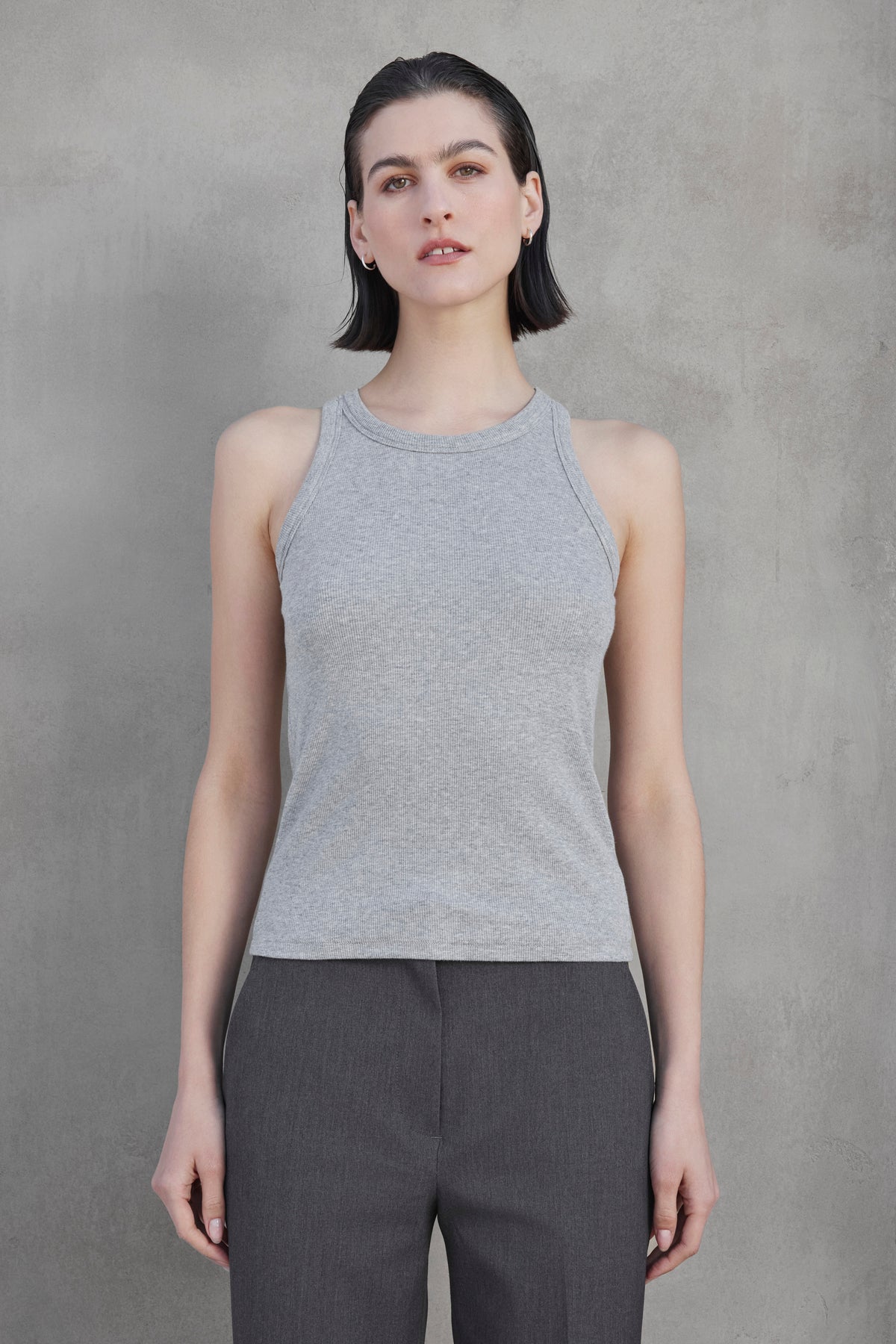   A person with short dark hair wears the versatile CRUZ TANK TOP from Velvet by Jenny Graham in a gray sleeveless top and dark gray pants, standing against a plain gray background. 