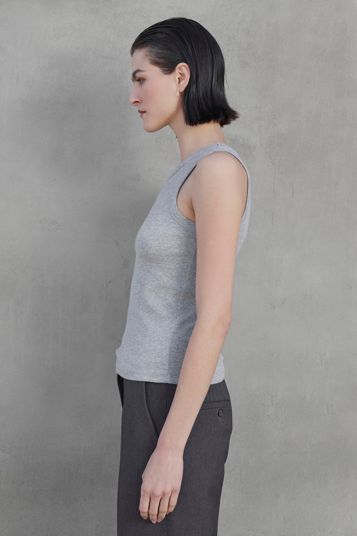   Side profile of a person with short dark hair, wearing the CRUZ TANK TOP from Velvet by Jenny Graham in a sleeveless gray ribbed cotton-modal blend shirt and dark gray pants, standing against a plain gray background. 