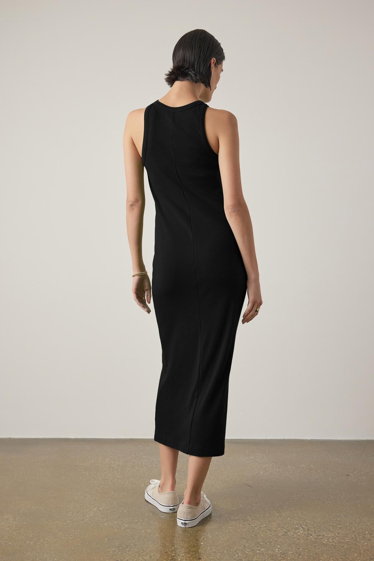   A woman in a sleek black sleeveless GRIFFITH DRESS from Velvet by Jenny Graham with a ribbed texture, seen from the back, paired with white sneakers, standing in a simple room with a concrete floor. 