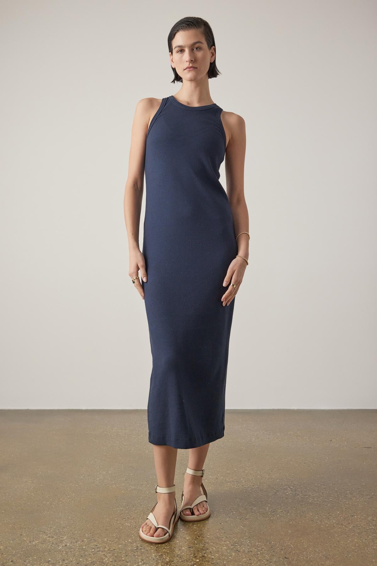   A woman in a Griffith dress by Velvet by Jenny Graham, a navy blue sleeveless midi dress with a crew neckline, stands in a neutral studio setting, paired with beige sandals. 