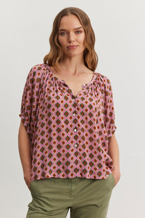 A woman with wavy hair wears the HARA TOP by Velvet by Graham & Spencer alongside green pants, standing with hands in pockets against a plain background.