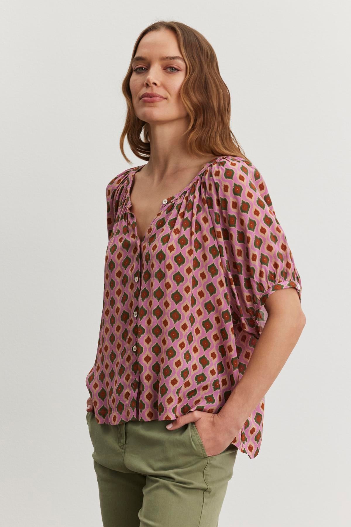   A person with long hair wears the HARA TOP by Velvet by Graham & Spencer and green pants, standing with hands in pockets against a plain background. 