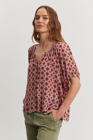 A person with long hair wears the HARA TOP by Velvet by Graham & Spencer and green pants, standing with hands in pockets against a plain background.