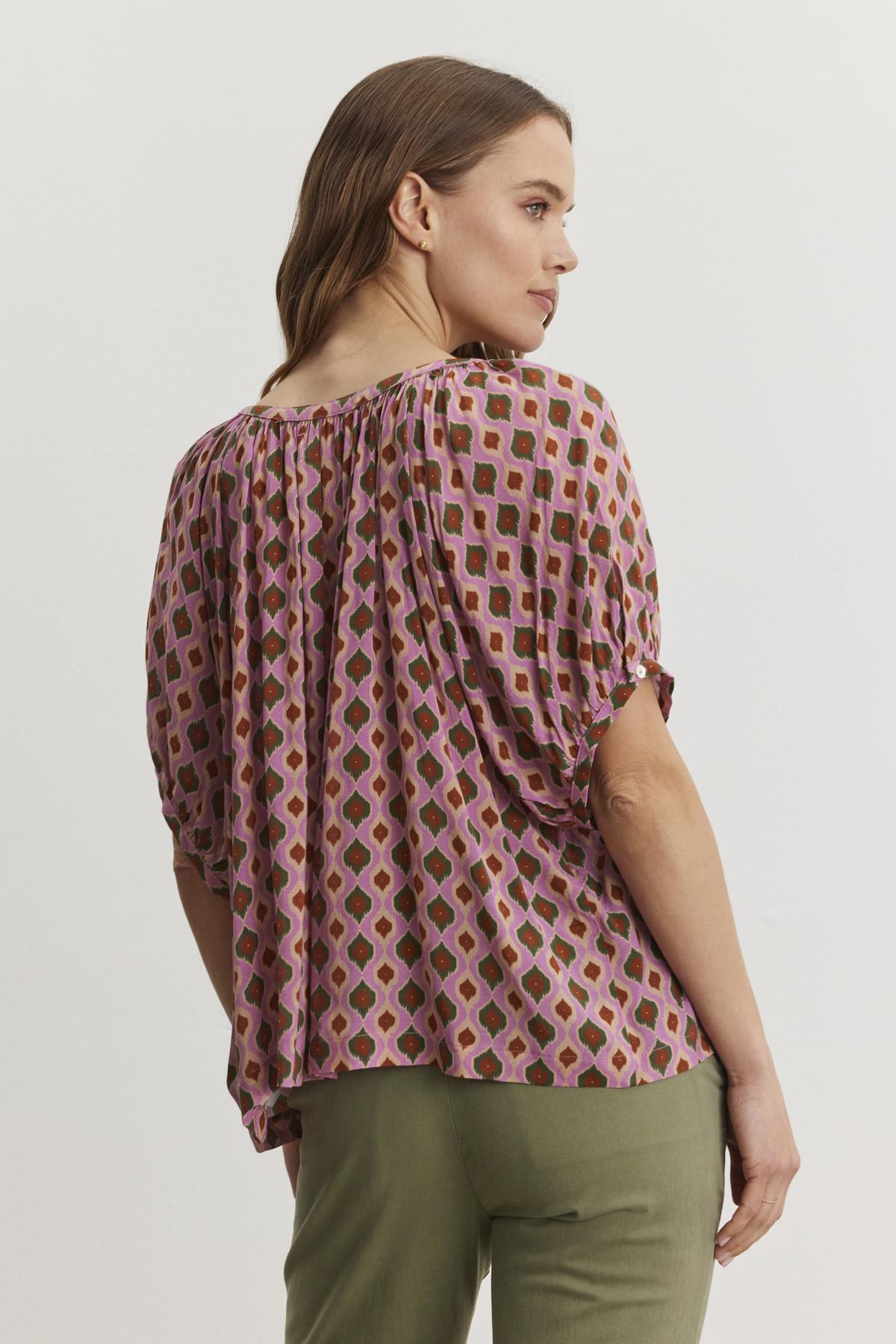 A woman with long hair is wearing the HARA TOP from Velvet by Graham & Spencer, which features a Moroccan print with pink and green shapes, paired with green pants. She is facing away, looking to the side.-38483587137729