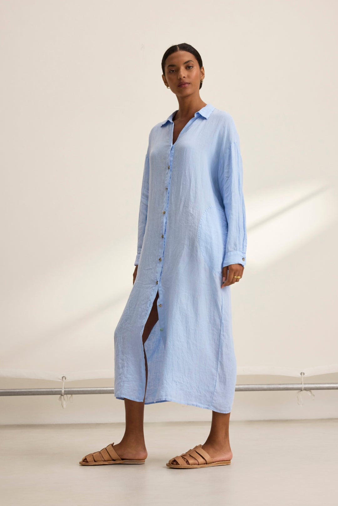   A person in the versatile, breathable NICOLE LINEN DRESS by Velvet by Graham & Spencer, styled with brown sandals, stands on a light floor against a plain backdrop. 