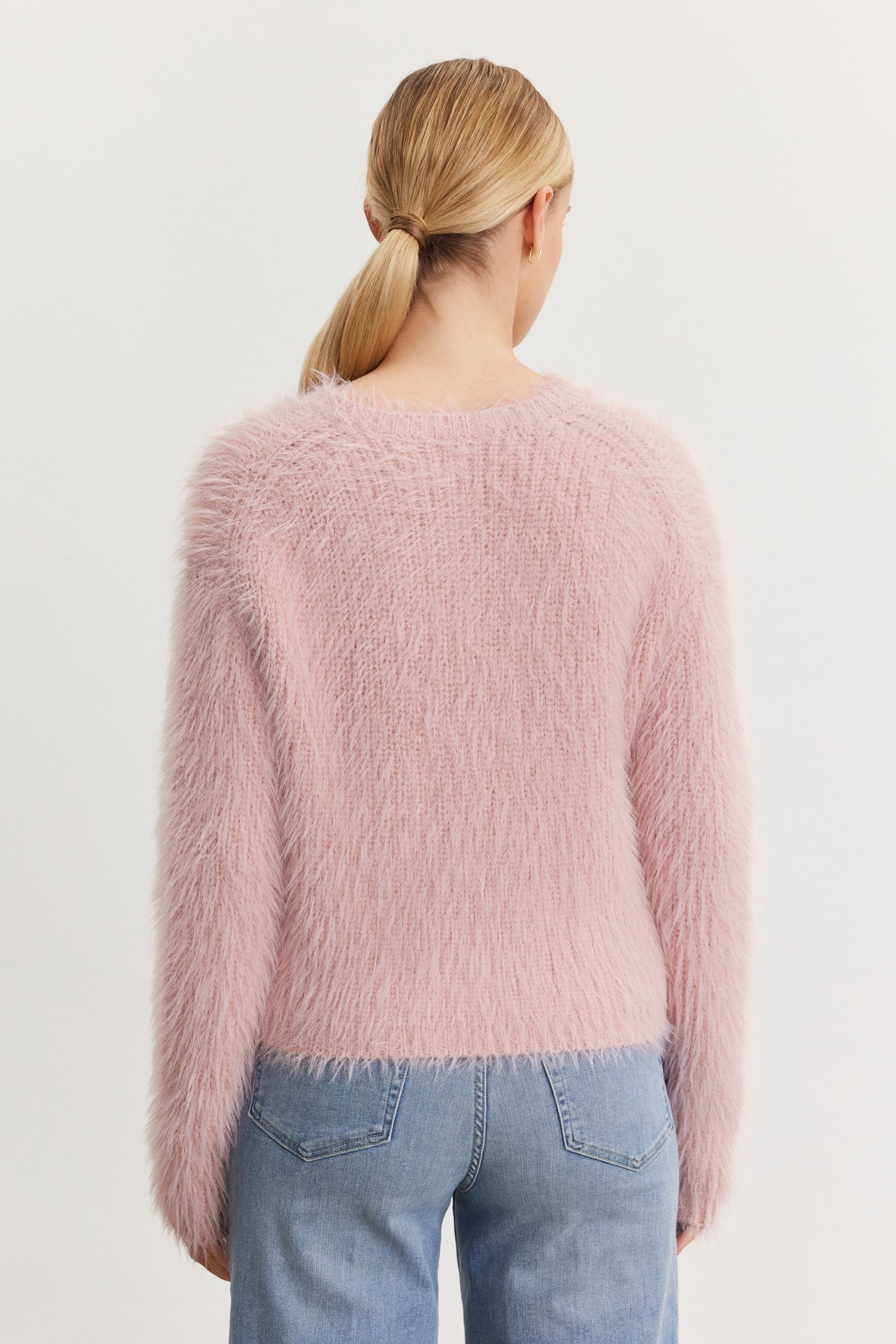 A person with blonde hair in a low ponytail is wearing the MAYLIE FEATHER YARN SWEATER by Velvet by Graham & Spencer, which features a fuzzy pink texture and crew neckline, along with blue jeans, while facing away from the camera.-38483622985921