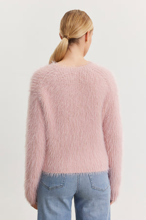 A person with blonde hair in a low ponytail is wearing the MAYLIE FEATHER YARN SWEATER by Velvet by Graham & Spencer, which features a fuzzy pink texture and crew neckline, along with blue jeans, while facing away from the camera.