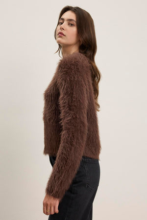 A person poses sideways in front of a plain background, wearing the MAYLIE FEATHER YARN SWEATER by Velvet by Graham & Spencer—a brown, furry long-sleeve top with a crew neckline—paired with black pants.