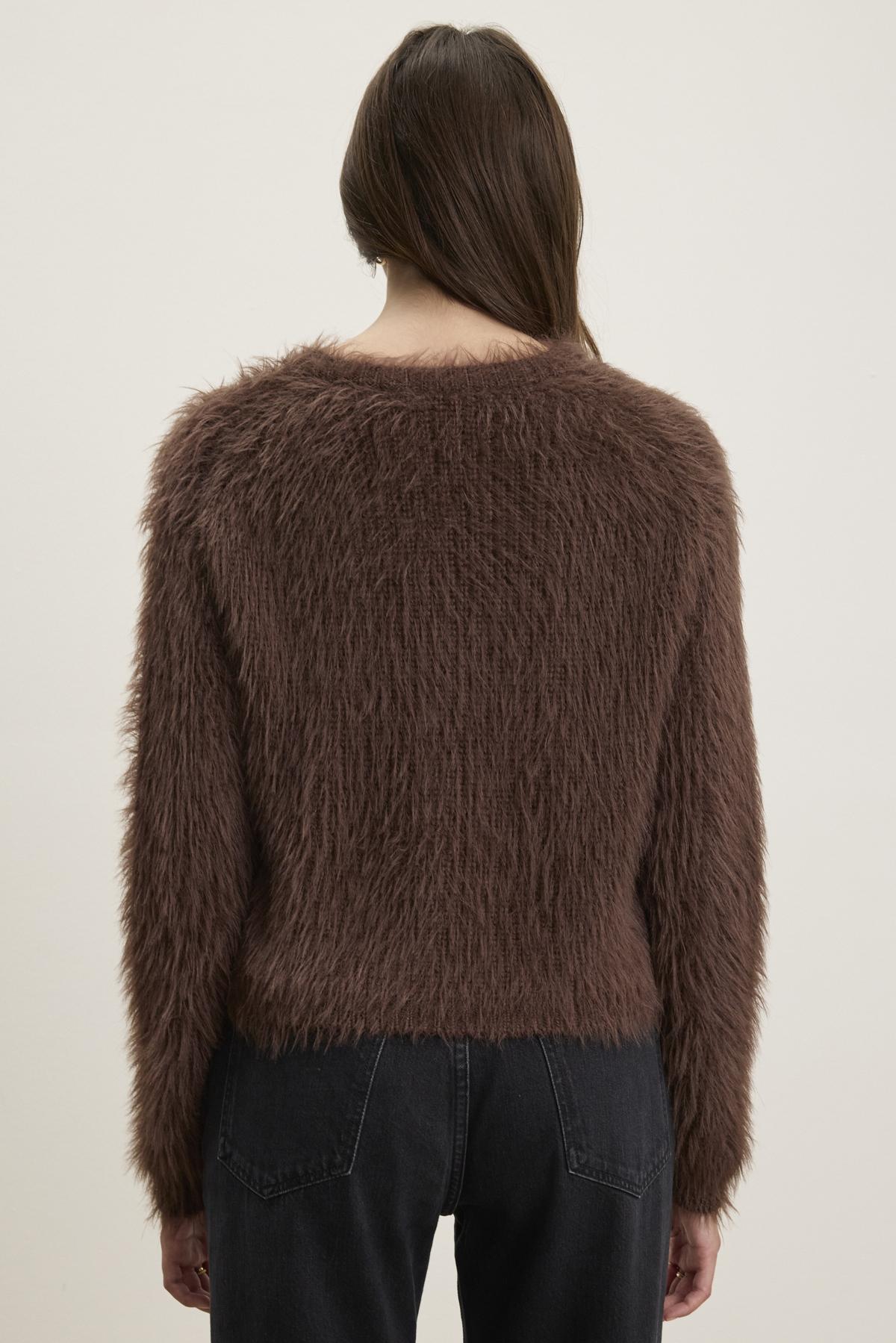   A person is seen from behind wearing the MAYLIE FEATHER YARN SWEATER from Velvet by Graham & Spencer, characterized by its brown feather yarn material and crew neckline, paired with dark jeans against a light background. 