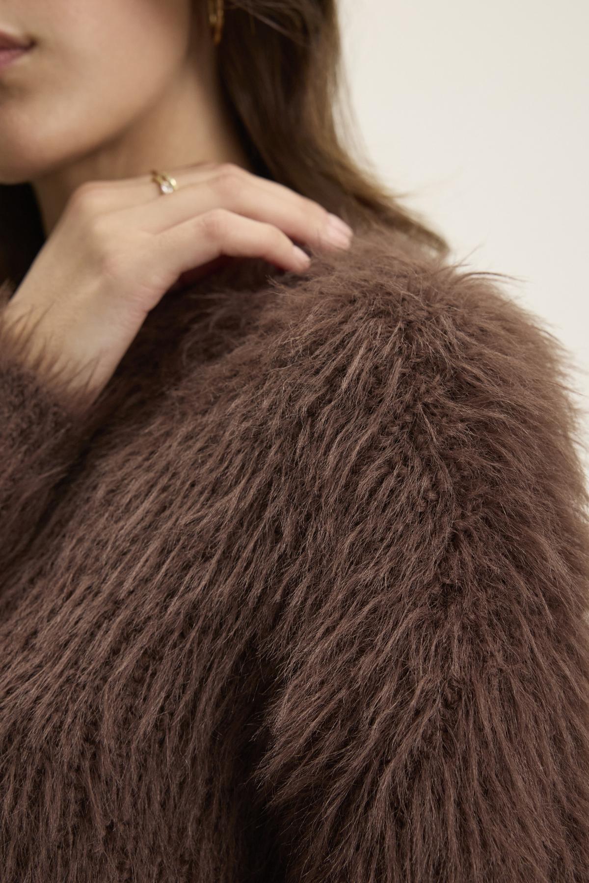   Close-up of a person touching their shoulder while wearing a fuzzy brown coat, with a glimpse of the MAYLIE FEATHER YARN SWEATER by Velvet by Graham & Spencer peeking out from underneath. 