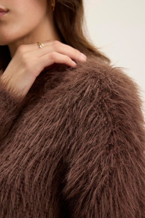 Close-up of a person touching their shoulder while wearing a fuzzy brown coat, with a glimpse of the MAYLIE FEATHER YARN SWEATER by Velvet by Graham & Spencer peeking out from underneath.