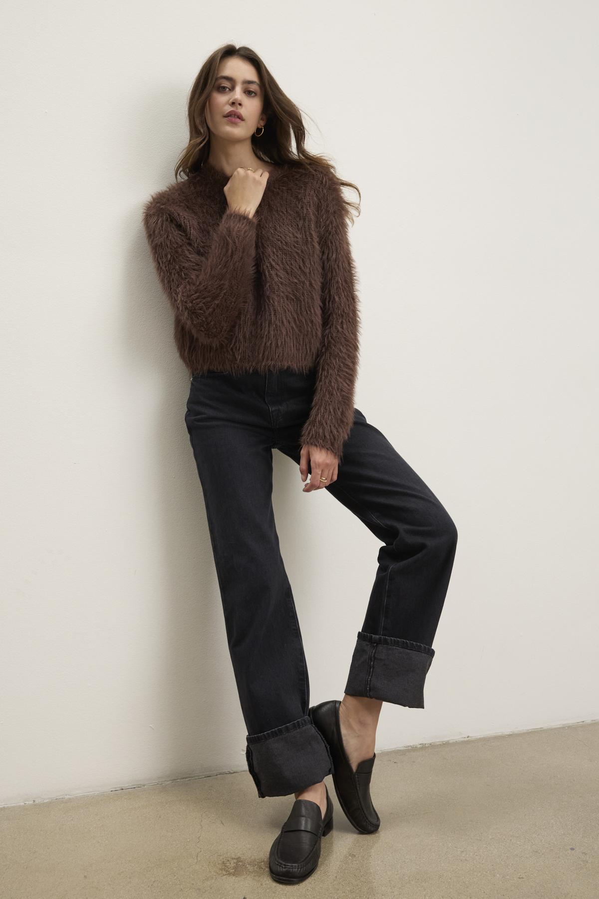   Wearing the MAYLIE FEATHER YARN SWEATER by Velvet by Graham & Spencer, a person poses against the stark white wall, dressed in dark rolled-up jeans and black shoes. 