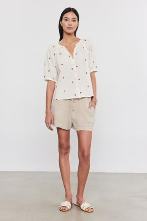 A woman standing in a studio, wearing a white polka-dot Amira top with floral embroidery, beige shorts, and white sandals by Velvet by Graham & Spencer.
