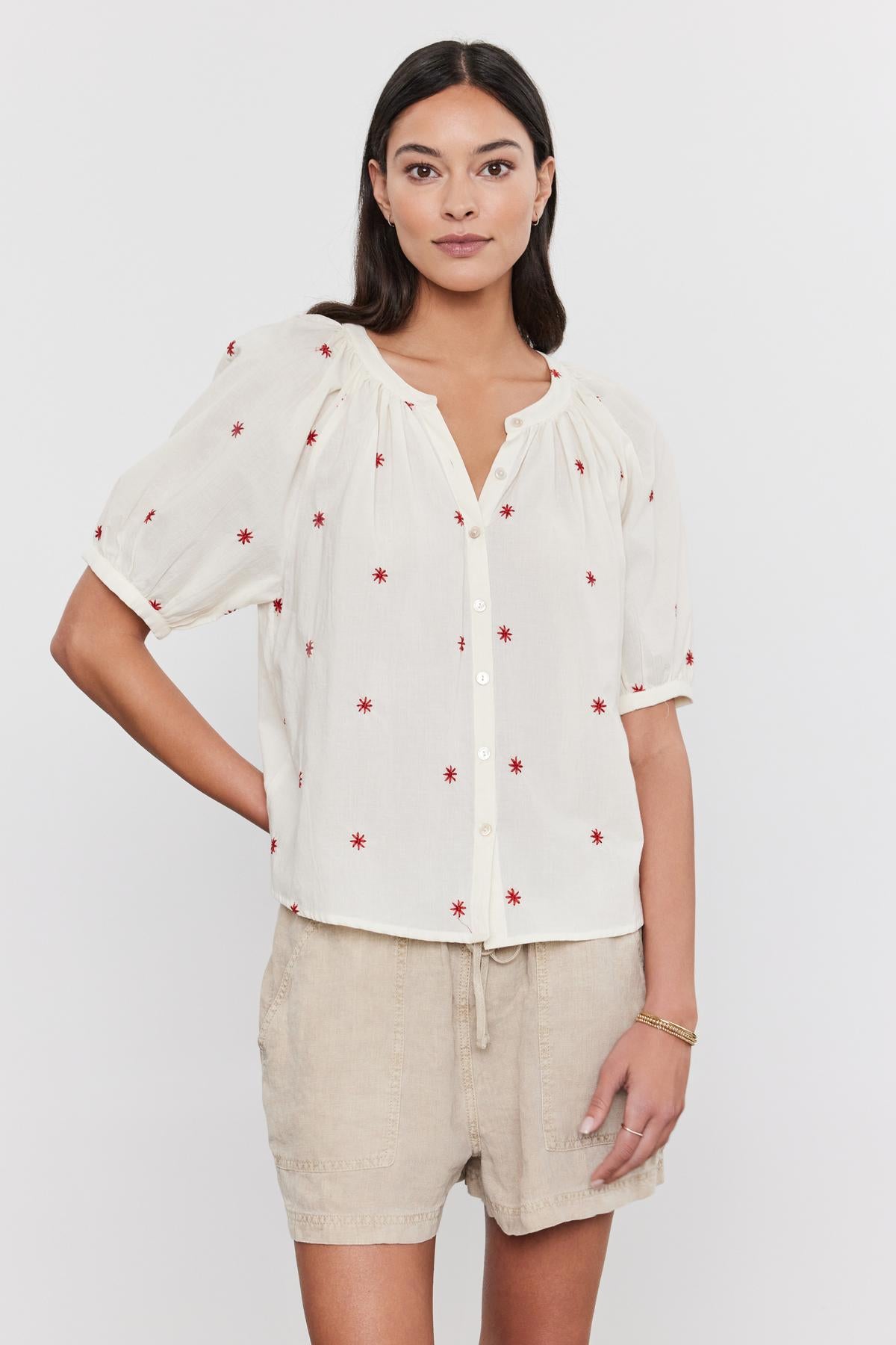   A woman wearing a white AMIRA TOP with red floral embroidery and beige shorts, standing against a plain background. Brand Name: Velvet by Graham & Spencer 