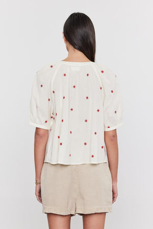 A woman from behind wearing a white Velvet by Graham & Spencer AMIRA TOP with red floral embroidery and button front detail, paired with a beige skirt.
