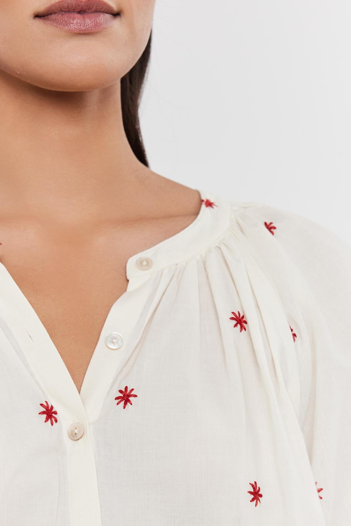   Close-up of a woman wearing the AMIRA TOP by Velvet by Graham & Spencer, a white blouse with red floral embroidery around the split neck. 