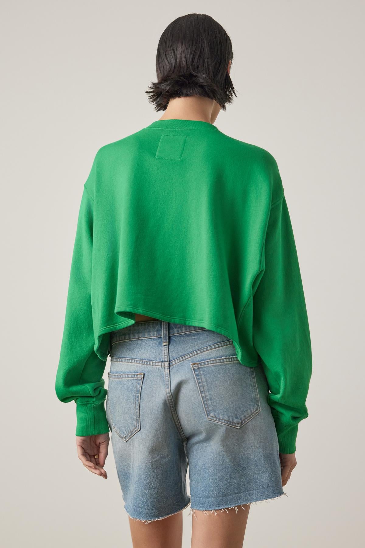   Rear view of a woman in a Velvet by Jenny Graham MALIBU SWEATSHIRT and denim shorts, standing against a beige background. 