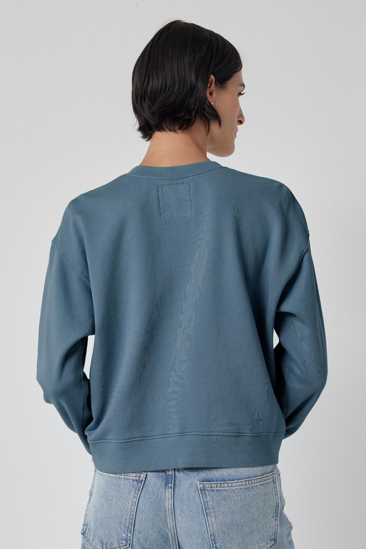   The back view of a woman wearing a blue Ynez Sweatshirt by Velvet by Jenny Graham with a dropped shoulder. 