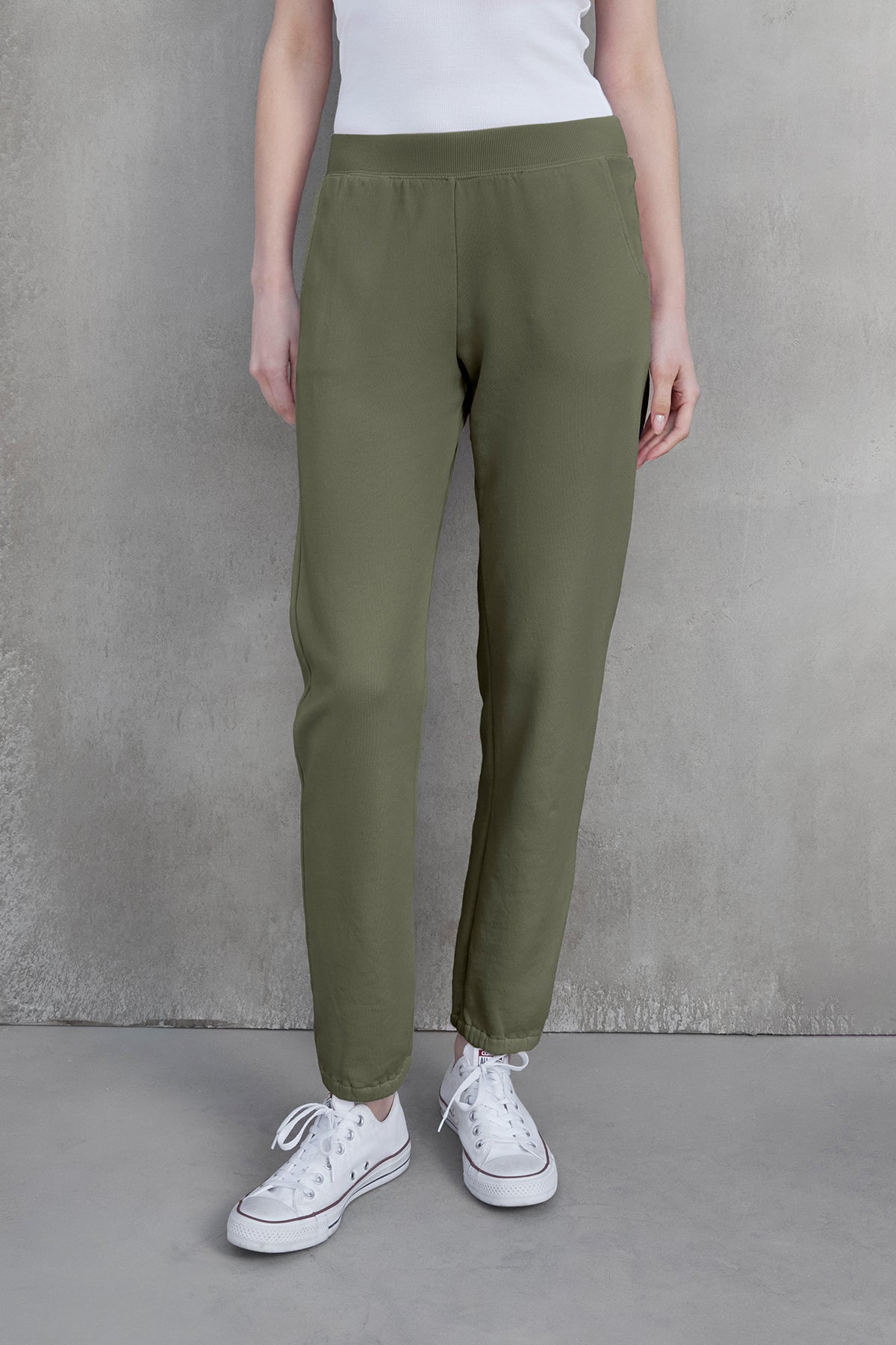   A person wearing ZUMA SWEATPANT in olive green by Velvet by Jenny Graham, paired with a white tank top and white sneakers, stands against a gray textured background, embracing sustainable fashion. 