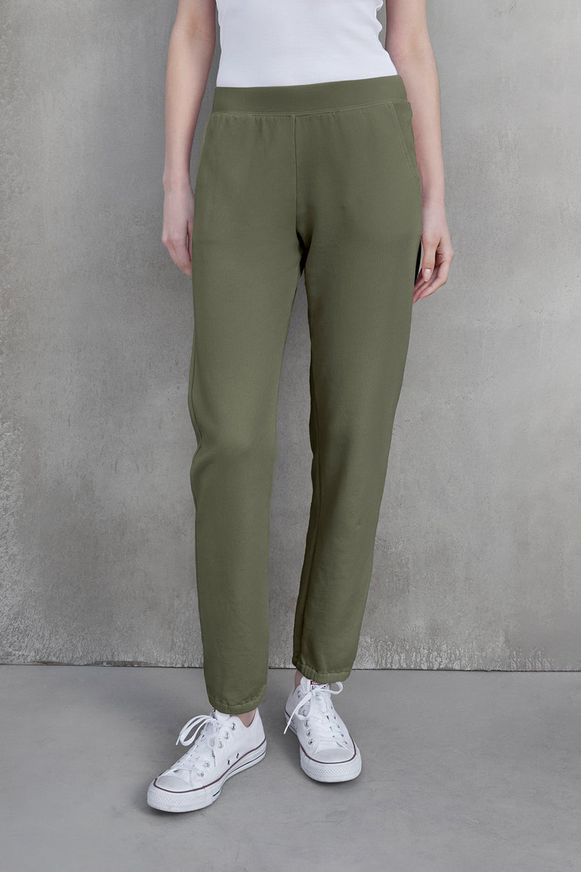 A person wearing ZUMA SWEATPANT in olive green by Velvet by Jenny Graham, paired with a white tank top and white sneakers, stands against a gray textured background, embracing sustainable fashion.