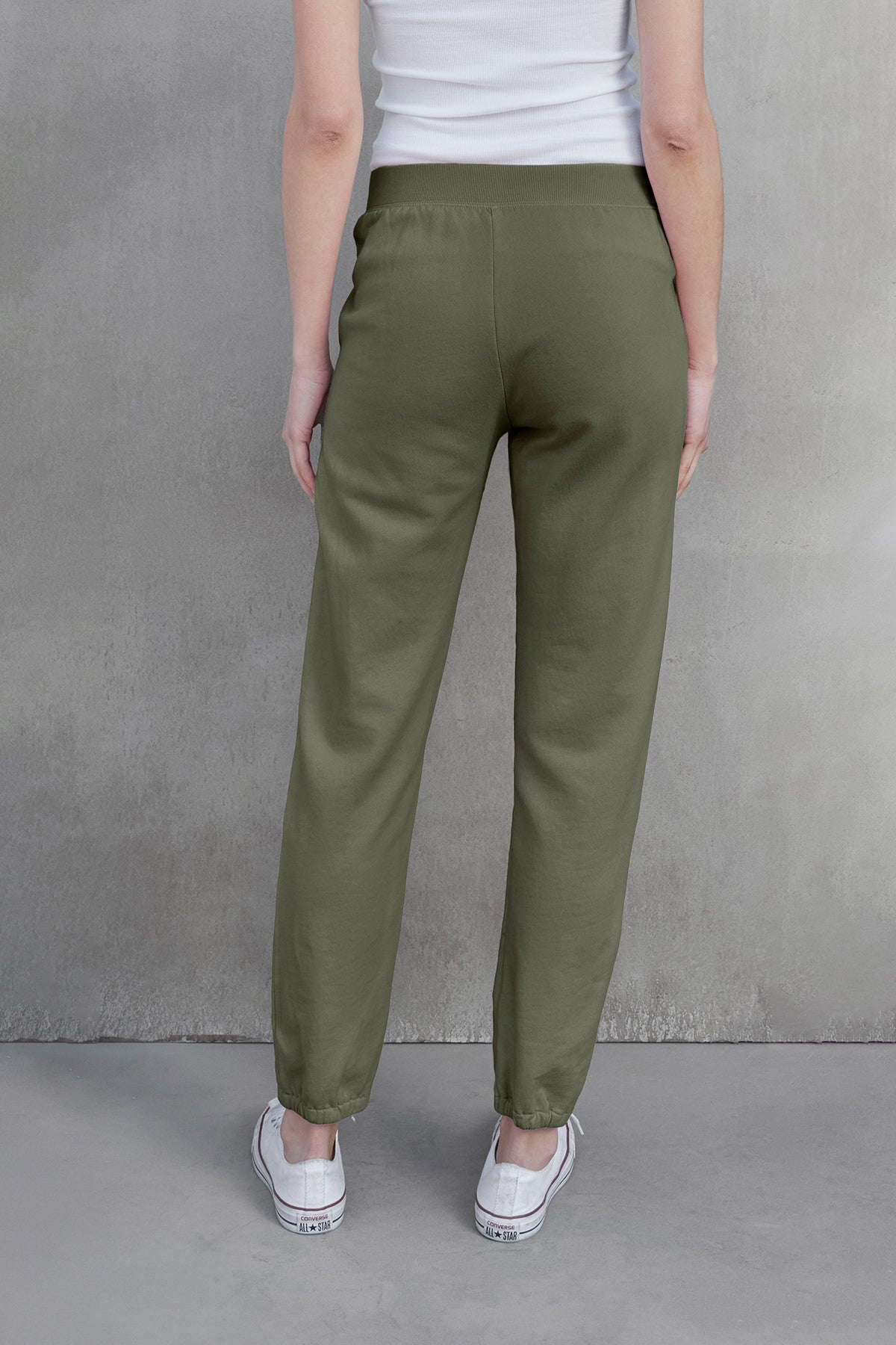   Against a gray backdrop, a person in velvet by Jenny Graham's ZUMA SWEATPANT, made of olive green organic cotton, pairs their look with a white top and sneakers. Only their back is visible, exuding an effortless mix of comfort and eco-conscious style. 