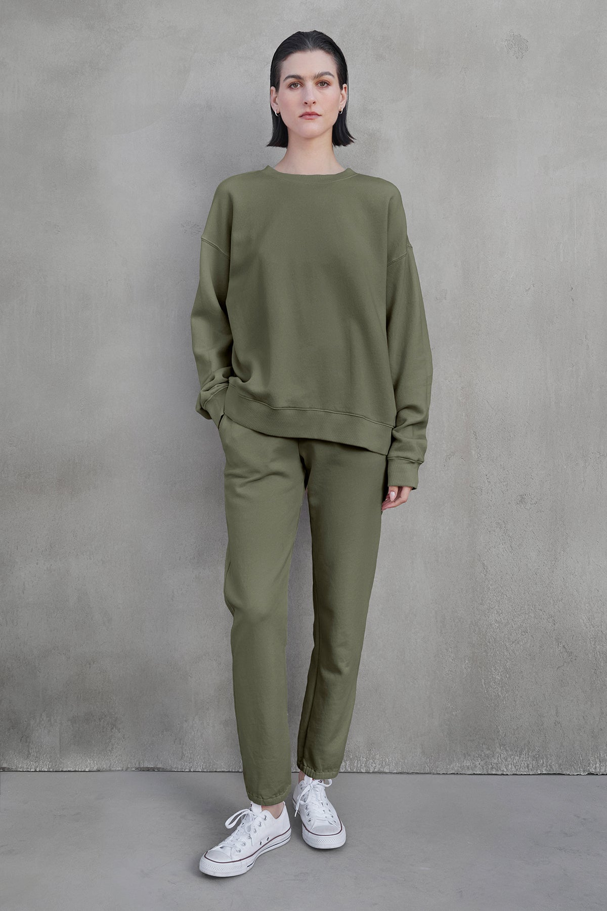   A person stands against a gray wall, wearing the ZUMA SWEATPANT and a matching sweatshirt in olive green from Velvet by Jenny Graham, paired with white sneakers, showcasing eco-friendly clothing. 