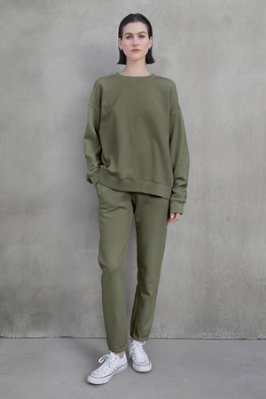 A person stands against a gray wall, wearing the ZUMA SWEATPANT and a matching sweatshirt in olive green from Velvet by Jenny Graham, paired with white sneakers, showcasing eco-friendly clothing.