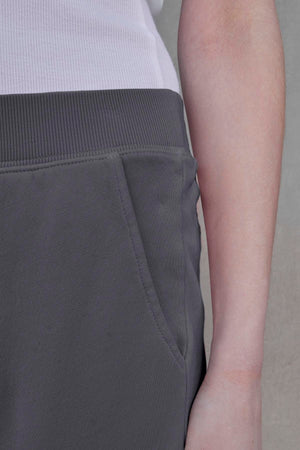 The image shows a close-up of a person wearing a white shirt and the ZUMA SWEATPANT by Velvet by Jenny Graham, made from dark gray organic cotton, with their hand resting in the pants pocket.