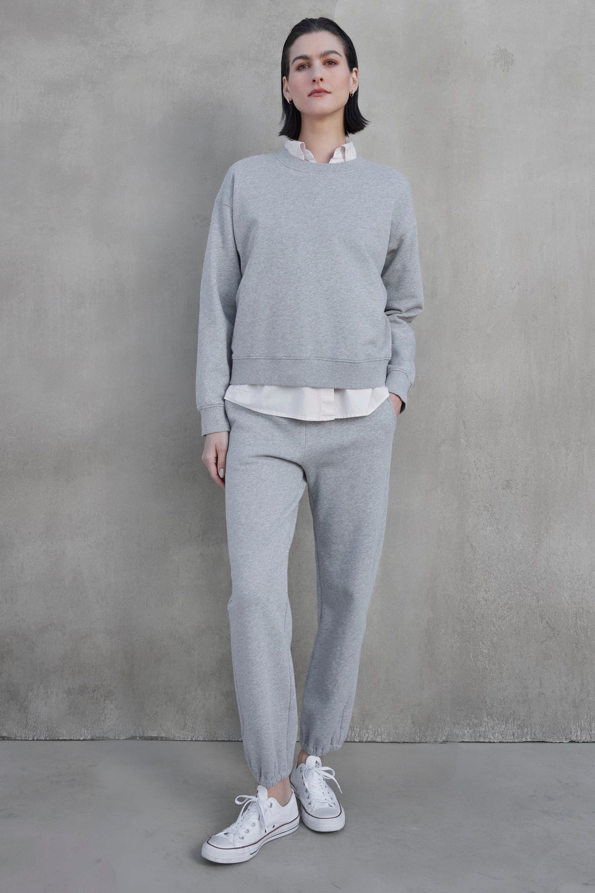   A person stands against a concrete wall wearing a grey sweatshirt and matching ZUMA SWEATPANTS from Velvet by Jenny Graham, made from organic cotton, layered over a white shirt, paired with white sneakers. 