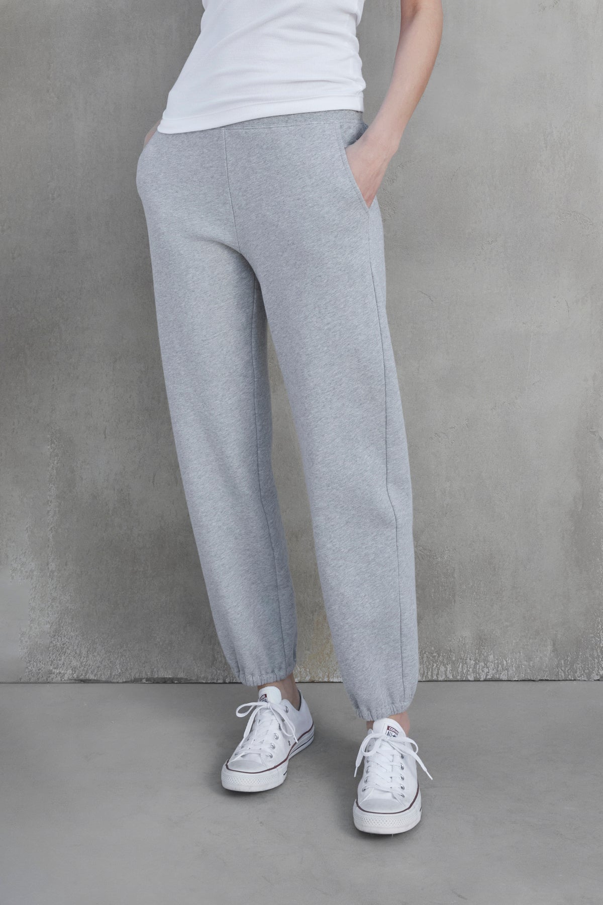 Person wearing a white t-shirt, ZUMA SWEATPANT gray joggers made of organic cotton by Velvet by Jenny Graham with hands in pockets, and white sneakers, standing against a gray concrete wall.-37659947794625